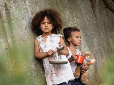 Selective focus of poor afrian american kid holding metal spoon and plate near brother with cup and